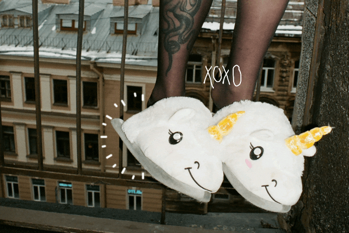GIF of a womans tattooed legs, she is wearing unicorn slippers and sitting on a balcony railing and you can see the houses from across the street.